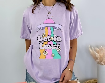 May include: A purple t-shirt with a graphic of a flying saucer with the text "Get in Loser" and rainbow stripes with flowers.