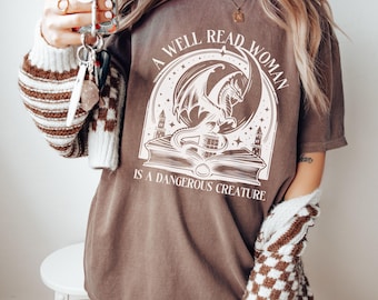 May include: A brown t-shirt with a white graphic of a dragon on a book. The graphic says "A well read woman is a dangerous creature."