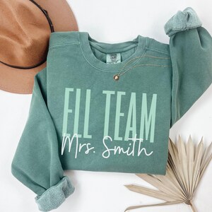 May include: A sage green sweatshirt with the text "FILL TEAM Mrs. Smith" in white and light green. A gold necklace with a small pendant is visible. A brown hat is in the background.
