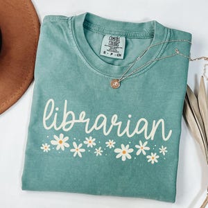 May include: A teal blue t-shirt with the word "librarian" in white script with a row of white daisies with yellow centers below the word.