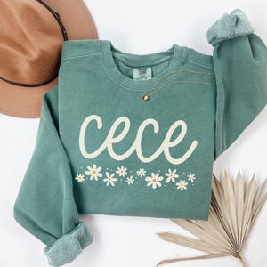 May include: A teal sweatshirt with the name "cece" in a white script font, and a row of white daisies with yellow centers. The sweatshirt has rolled-up sleeves and a crew neck.