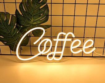 May include: White neon sign with the word "Coffee" in a cursive font. The sign is illuminated and is hanging against a black wire mesh background.