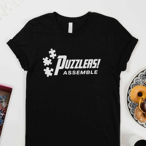 May include: Black t-shirt with the white text "Puzzlers! Assemble" and puzzle piece graphics. A plate of doughnuts and a coffee cup are also visible in the image.