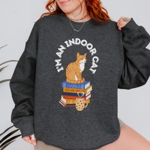 May include: A dark grey sweatshirt with the text "I'm an indoor cat" in a circle around a cartoon orange cat sitting on a stack of books with a coffee mug on top.