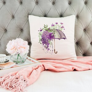 Może przedstawiać: Biała poduszka z akwarelowym wzorem fioletowego parasola z fioletowymi kwiatami i zielonymi liśćmi. Poduszka leży na szarym, pikowanym zagł&oacute;wku z r&oacute;żowym kocem.