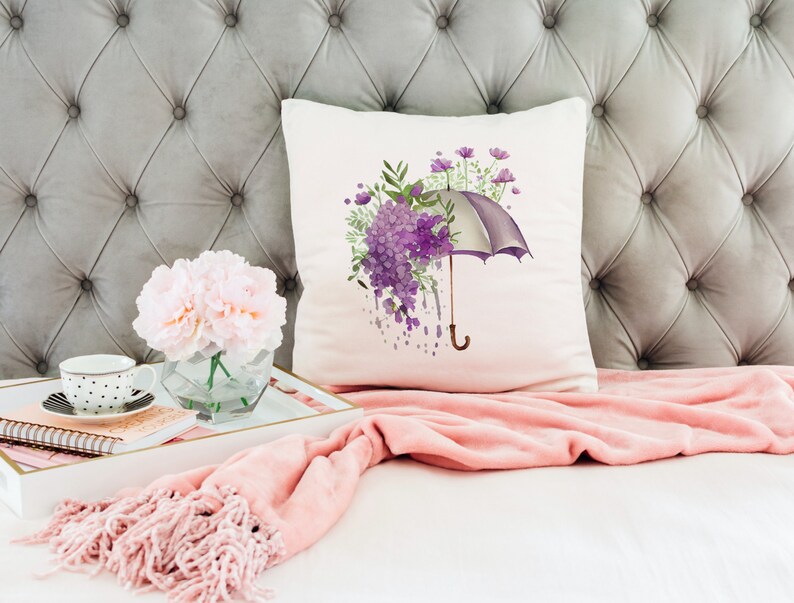 Może przedstawiać: Biała poduszka z akwarelowym wzorem fioletowego parasola z fioletowymi kwiatami i zielonymi liśćmi. Poduszka leży na szarym, pikowanym zagł&oacute;wku z r&oacute;żowym kocem.