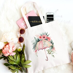 Może przedstawiać: Biała torba typu tote z pł&oacute;tna z r&oacute;żowym i zielonym wzorem kwiatowym z r&oacute;żowym parasolem. Torba jest wypełniona r&oacute;żowym notesem, r&oacute;żową książką, okularami przeciwsłonecznymi i telefonem kom&oacute;rkowym.