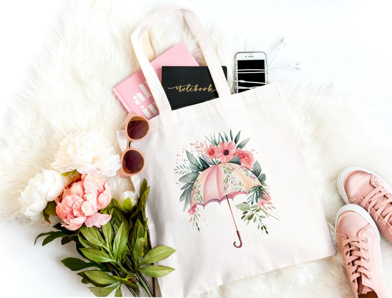 Może przedstawiać: Biała torba typu tote z pł&oacute;tna z r&oacute;żowym i zielonym wzorem kwiatowym z r&oacute;żowym parasolem. Torba jest wypełniona r&oacute;żowym notesem, r&oacute;żową książką, okularami przeciwsłonecznymi i telefonem kom&oacute;rkowym.