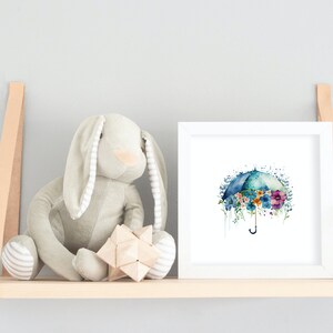 Może przedstawiać: Biały, oprawiony wydruk akwareli przedstawiający niebieski parasol z kolorowymi kwiatami. Parasol otoczony jest jasnoniebieskim akwarelowym rozmyciem. Wydruk znajduje się na p&oacute;łce z pluszowym kr&oacute;likiem i drewnianą zabawką.