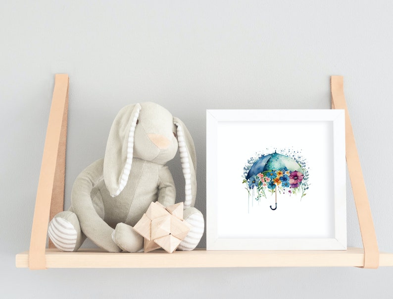 Może przedstawiać: Biały, oprawiony wydruk akwareli przedstawiający niebieski parasol z kolorowymi kwiatami. Parasol otoczony jest jasnoniebieskim akwarelowym rozmyciem. Wydruk znajduje się na p&oacute;łce z pluszowym kr&oacute;likiem i drewnianą zabawką.