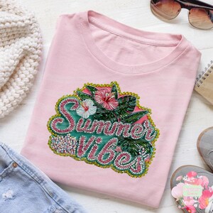 Op de afbeelding: Een lichtroze T-shirt met een kleurrijke "Summer Vibes" print. Het ontwerp bevat tropische bloemen, bladeren en glitter. Het T-shirt wordt vergezeld door een zonnebril, een notitieblok en een paar schoenen. Thema: zomer mode.