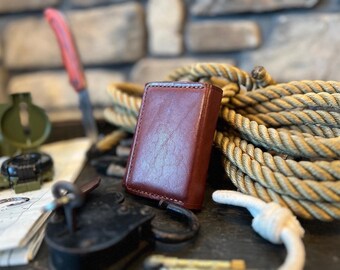 May include: A brown leather wallet with a textured surface sits on a table with a compass, rope, and a lock.