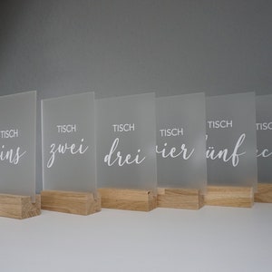 Könnte beinhalten: Ein Set aus sechs Tischkarten aus mattiertem Acryl mit Holzsockeln. Jedes Schild trägt das Wort "Tisch" über einer elegant geschriebenen Nummer. Die Nummern sind "eins", "zwei", "drei", "vier", "fünf" und "sechs".