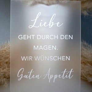 Könnte beinhalten: Ein mattes Acrylschild mit dem Text "Liebe geht durch den Magen. Wir wünschen Guten Appetit" in weißer, kursiv geschriebener Schrift.