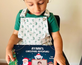 May include: A child in a green top and white patterned dungarees holds a book titled "Ayaan's Christmas Adventure!". The book cover displays illustrations of Santa Claus, an elf, and a Christmas tree.