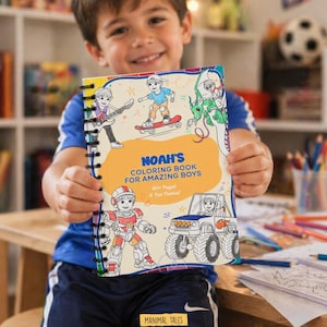 May include: A spiral-bound coloring book titled "Noah's Coloring Book for Amazing Boys" is held by a smiling child. The cover features illustrations of boys skateboarding, playing guitar, and riding a monster truck. The book has 40+ pages and 6 fun themes.