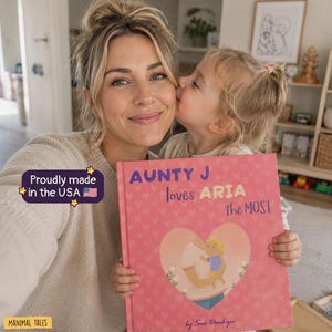 May include: A hardcover children's book with a pink cover featuring a heart-shaped illustration of a person holding a child. The text reads "AUNTY J loves ARIA the MOST" and "by Sue Donhija". A purple badge states "Proudly made in the USA".
