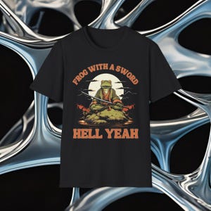 May include: Black t-shirt featuring a graphic of a frog with a sword, seated on a rock with a mountain and moon backdrop. The text "FROG WITH A SWORD" and "HELL YEAH" are in orange. The design has a vintage, illustrated style.