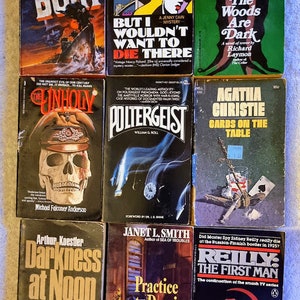 May include: A collection of vintage paperback books with various titles and authors. The covers feature illustrations and text, including titles like "The Night Boat," "Poltergeist," and "Darkness at Noon." The books are arranged on a light blue surface.