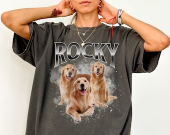 Op de afbeelding: Een donkergrijs T-shirt met een afbeelding van drie golden retriever honden en het woord "ROCKY" in een zilveren metallic lettertype.