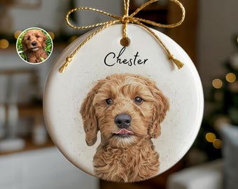 May include: A round, cream-colored Christmas ornament featuring a detailed portrait of a brown dog with its tongue sticking out. The name "Chester" is written above the dog's image in black script. The ornament is suspended by a gold string and bow.