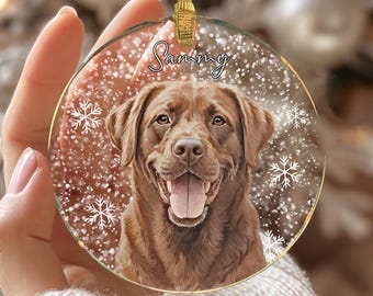 May include: A clear, round Christmas ornament featuring a smiling brown Labrador Retriever. The ornament has the name "Sammy" in cursive script and snowflake accents. A gold ribbon is attached for hanging.