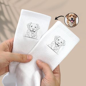 May include: A pair of white socks featuring a black line drawing of a dog on each sock. The dog is smiling with its tongue out. The socks are made from a soft, comfortable material and are ideal for everyday wear.