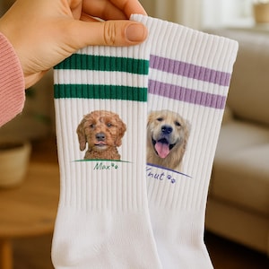 Könnte beinhalten: Weiße Sportsocken mit grünen und lila Streifen. Jede Socke zeigt ein Hundeporträt mit einem Namen darunter. Ein Hund ist ein Golden Doodle, der andere ein Golden Retriever. Die Socken werden von einer Person hochgehalten.