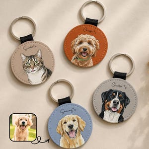 Può includere: Una collezione di portachiavi personalizzati per animali domestici con ritratti dipinti di un gatto e cani. Ogni portachiavi ha un design circolare in pelle con un anello di metallo e un cinturino in pelle nera. I nomi degli animali sono scritti sopra i loro ritratti.