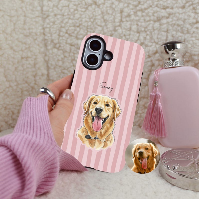 Op de afbeelding: Roze gestreepte telefoonhoesje met een illustratie van een Golden Retriever en de naam "Sammy". De hond steekt zijn tong uit. Een kleinere afbeelding van de hond staat onderaan. De hoes wordt getoond met een roze trui en parfumfles.