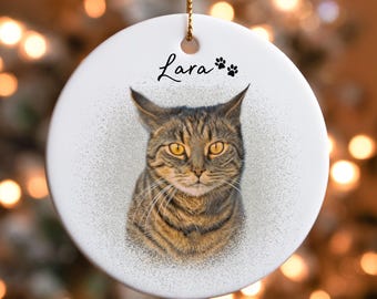 Op de afbeelding: Een witte, ronde keramische ornament met een gedetailleerd portret van een tabby kat met gele ogen. De naam van de kat, "Lara", is in cursief boven de afbeelding geschreven, vergezeld van pootafdrukken. Een gouden touwtje is bevestigd om op te hangen.