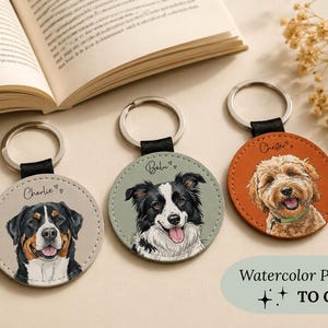 Könnte beinhalten: Drei runde Schlüsselanhänger mit Aquarell-Haustierporträts. Jeder Schlüsselanhänger zeigt eine andere Hunderasse: einen Berner Sennenhund, einen Border Collie und einen Goldendoodle. Die Schlüsselanhänger haben silberne Ringe und schwarze Lederriemen.