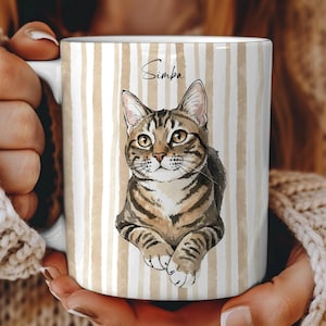Può includere: Tazza in ceramica bianca con un'illustrazione di un gatto tigrato e il nome "Simba". La tazza ha un motivo a righe verticali beige e bianco. Il gatto è marrone e beige.