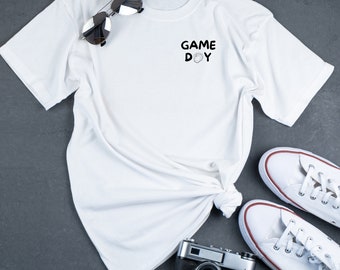May include: White t-shirt with the text "GAME DAY" and a hand graphic on the front. The shirt is laid out on a gray surface with a pair of white sneakers and a vintage camera.