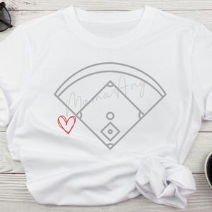 Puede incluir: Camiseta blanca con un contorno gris de un campo de béisbol y el texto "Mama's Angel" en cursiva. Un corazón rojo se encuentra en la esquina inferior izquierda.