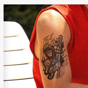 Puede incluir: Un diseño de tatuaje temporal con una ilustración en tinta negra de una figura similar a un mono que sostiene un bastón, rodeada de patrones de llamas estilizados. El diseño está aplicado en un brazo con una camiseta roja sin mangas.