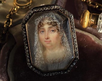 Può includere: Una spilla ottagonale con un ritratto in miniatura di una persona con capelli castani che indossa un velo di pizzo bianco. Il ritratto è racchiuso in una cornice di metallo scuro bordata da piccole pietre chiare e scintillanti.
