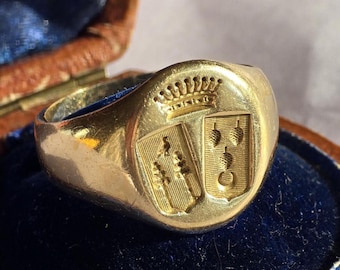 Può includere: Un anello sigillo in oro con uno stemma inciso raffigurante due scudi affiancati sormontati da una corona. Lo scudo sinistro mostra due simboli verticali a forma di albero, mentre quello destro presenta tre forme a pigna impilate sopra una luna.
