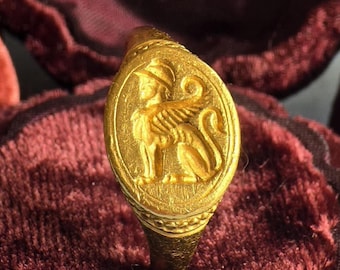 Può includere: Un anello in oro con una montatura ovale caratterizzata da un rilievo di una sfinge alata che indossa un elmo. La superficie dell'oro presenta una finitura opaca e testurizzata, con la figura centrale raffigurata di profilo verso sinistra.