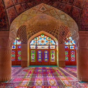 Pode incluir: Vista interior de uma mesquita com arquitetura ornamentada. O teto e as paredes apresentam padrões e desenhos intrincados. Vitrais em cores vibrantes projetam luz sobre os tapetes e colunas estampadas.
