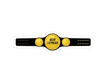 May include: A sticker featuring a championship belt design. The belt is black with gold accents, including three circular medallions. The central medallion reads "BEST CUTMAN" in bold, black text.