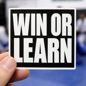 May include: A square sticker with the words "WIN OR LEARN" in bold white capital letters on a black background. The sticker is held by a hand, with a blurred background of a blue surface and people.