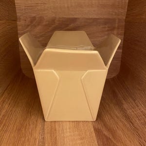 May include: A beige ceramic takeout container with a folded design. The container is open, revealing a glimpse of the contents inside. The container is sitting on a wooden surface.