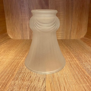 May include: A frosted glass lampshade with a flared base and a decorative, draped design around the top. The lampshade is a soft, off-white colour and is set against a wooden background.