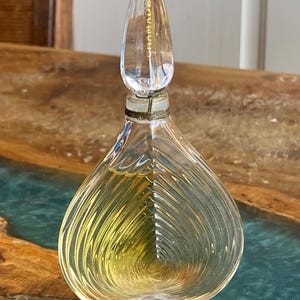 Könnte beinhalten: Ein klarer Glasparfümflakon mit tropfenförmigem Körper und spitzem Verschluss. Die Flasche enthält eine goldfarbene Flüssigkeit und das Wort "CHAMADE" ist auf dem Verschluss aufgedruckt. Der Sockel ist rechteckig.