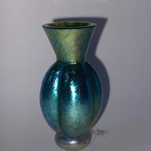 Puede incluir: Un pequeño jarrón de vidrio iridiscente con un cuerpo bulboso y un cuello acampanado. El jarrón tiene una combinación de colores verde azulado y azul con una textura moteada y una base plateada. El jarrón mide unos 13 cm de altura.