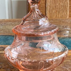 Peut inclure: Un pot en verre rose avec un couvercle. Le pot présente des motifs floraux gravés et un bouton à facettes. Le pot est en verre rose et a une base ronde. Le couvercle s'adapte parfaitement sur le dessus du pot.