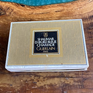 May include: A rectangular box with a gold and white design. The box features a gold square with the text "SHALIMAR L'HEURE BLEUE CHAMADE GUERLAIN PARIS" in black and gold. The box is on a wooden surface.