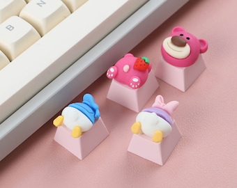 May include: Four pink keycaps with cartoon character designs. The keycaps feature a bear, a duck, a pig, and a strawberry character. The keycaps are on a pink surface.