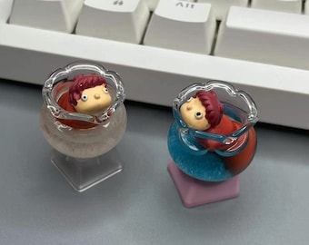 May include: Two small glass figurines of people with red hair, each sitting in a clear glass bowl. One bowl is filled with a red liquid and the other with a blue liquid.